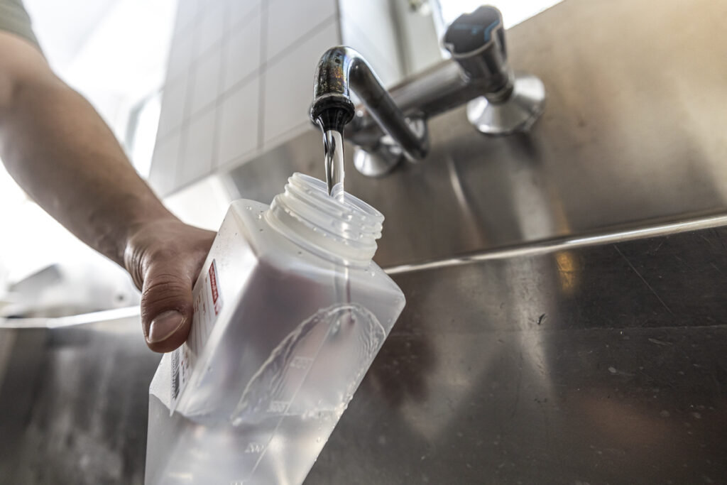 Hand füllt sterile Probenflasche direkt am Wasserhahn zur mikrobiologischen Untersuchung der Trinkwasserinstallation.