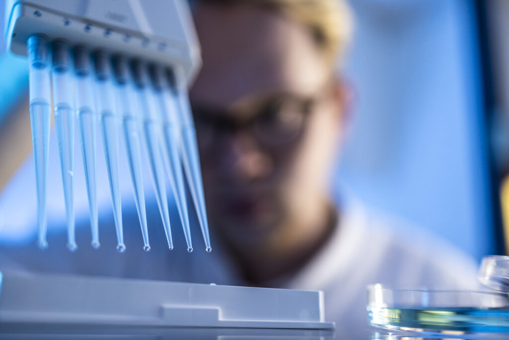 Mehrkanalpipette mit Tropfen über Mikrotiterplatte; unscharfe Laborfachkraft und Petrischale im Hintergrund.